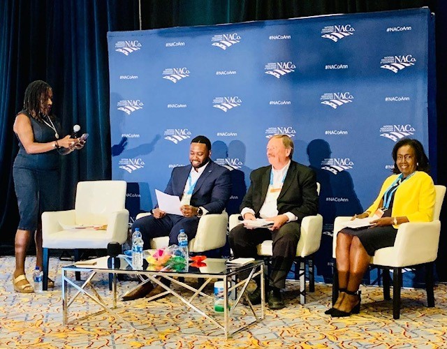 A group of people, including BOCC Chair Brenda Howerton, sit on stage at the NACo annual convention.